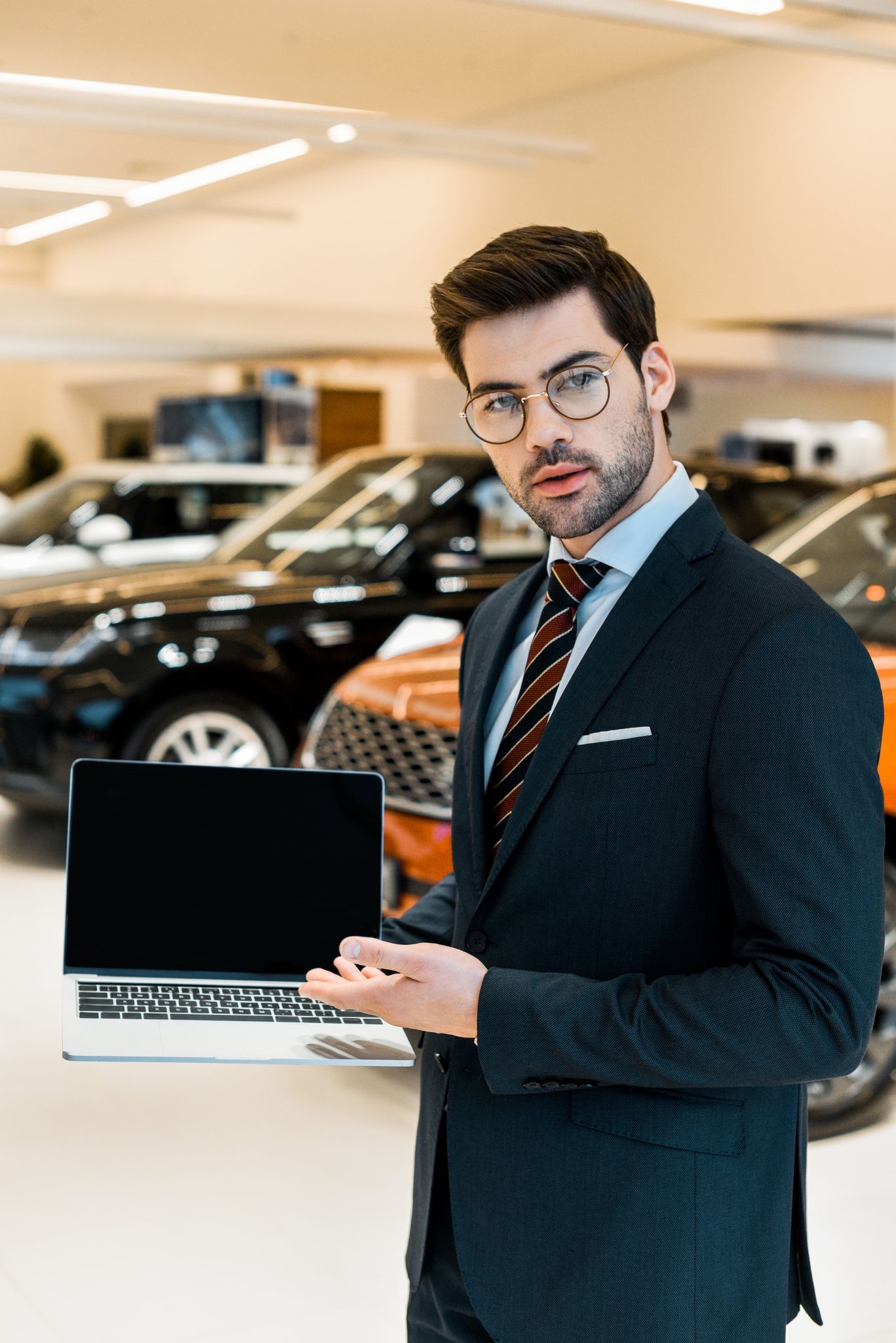 young male car dealer in eyeglasses pointing at laptop with blank screen in car salon