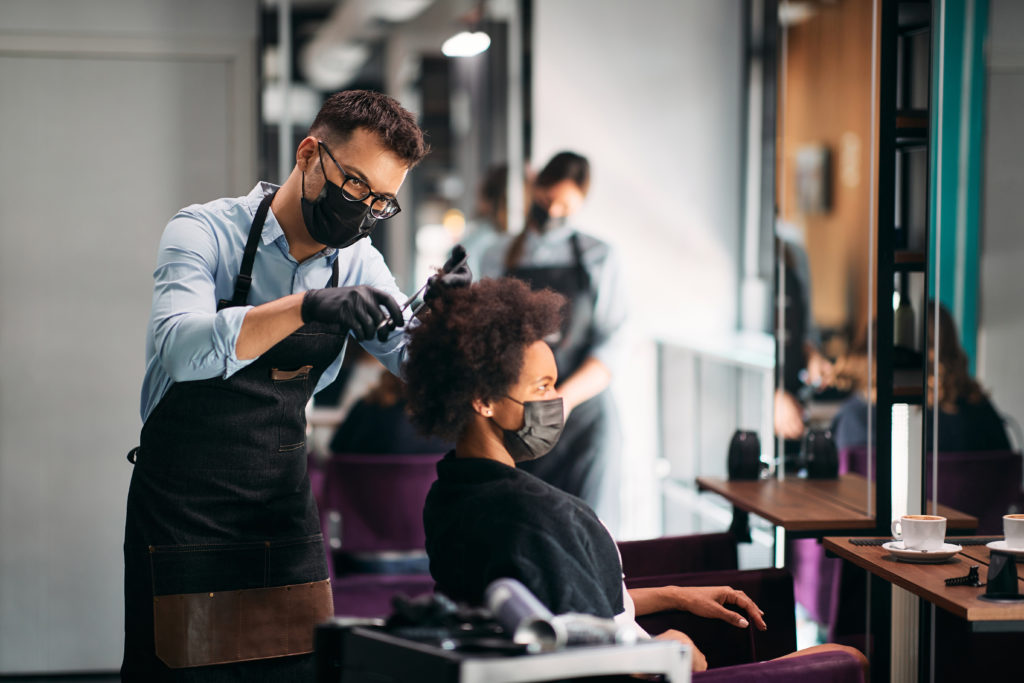 Hairdresser and his African American client wearing face masks during haircut at the salon.