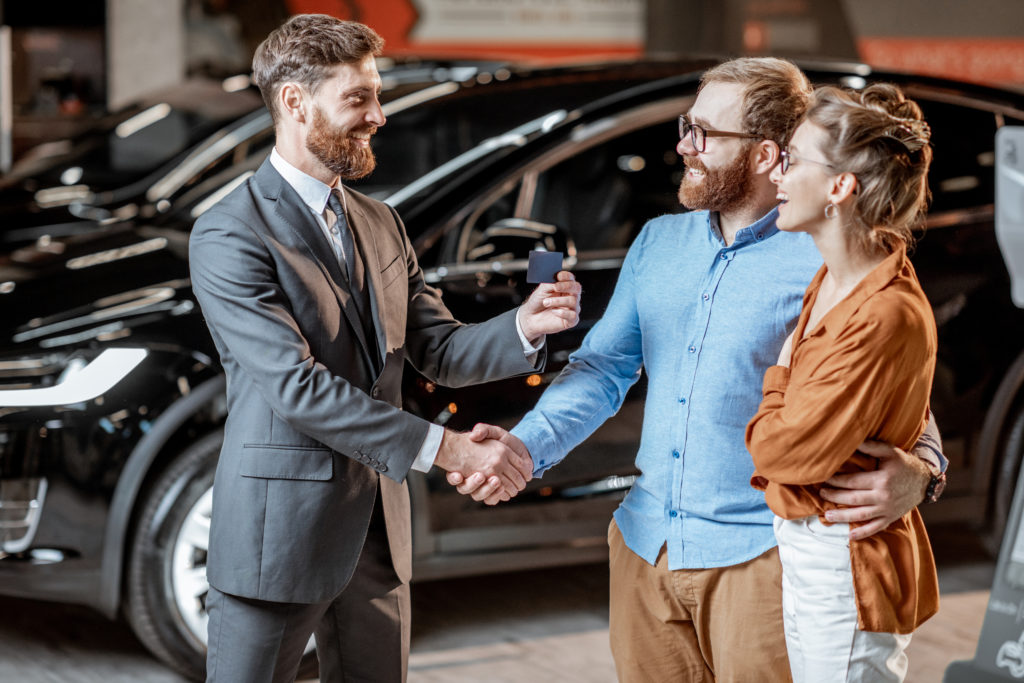 Sales manager giving a card key for a young couple, buying new electric car at the modern showroom of the car dealership