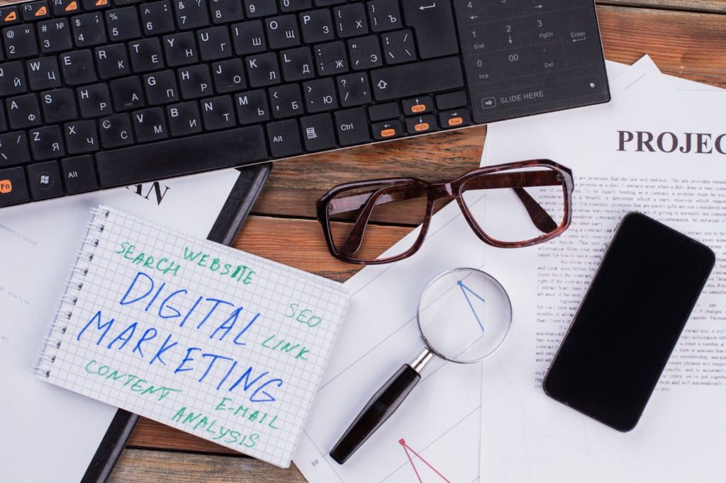 A desk with a keyboard, smartphone, glasses, magnifying glass, and a notebook labeled "digital marketing," surrounded by various documents.