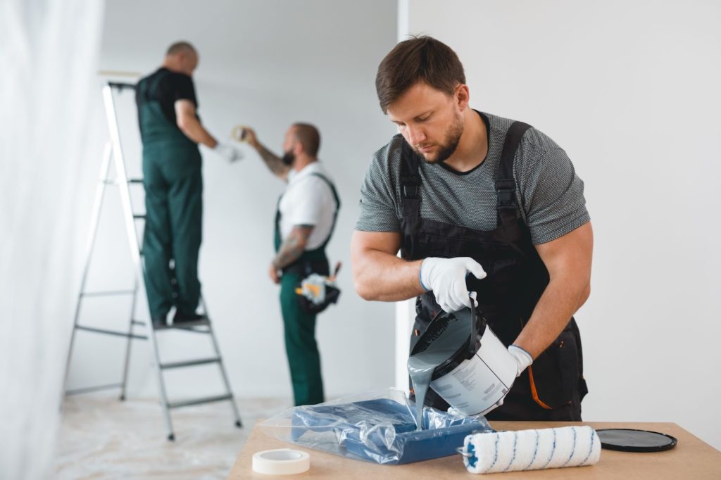 A man in overalls painting a room with a roller brush, focusing on the details of the walls and furniture.