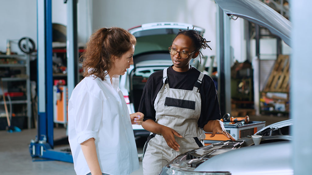 Friendly mechanic assisting customer with car maintenance in auto repair shop. Qualified specialist in garage looking over car components with woman, servicing her vehicle during annual checkup