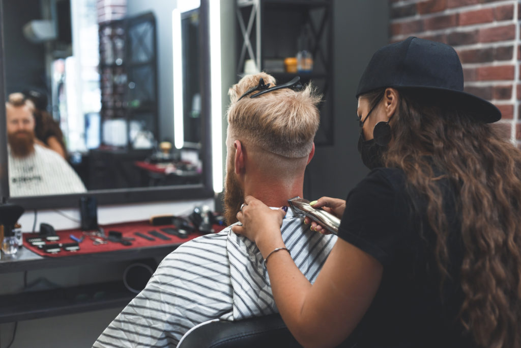 Female barber in mask cuts a man hairs with hair clipper. Hairstyle during social distancing.