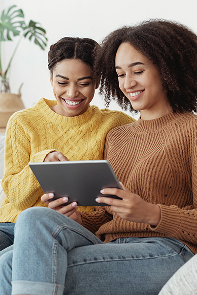 Enjoying weekend and leisure time together or shopping online at home. Cheerful young attractive african american sisters watch at tablet and choose movie or purchases on sofa in living room interior