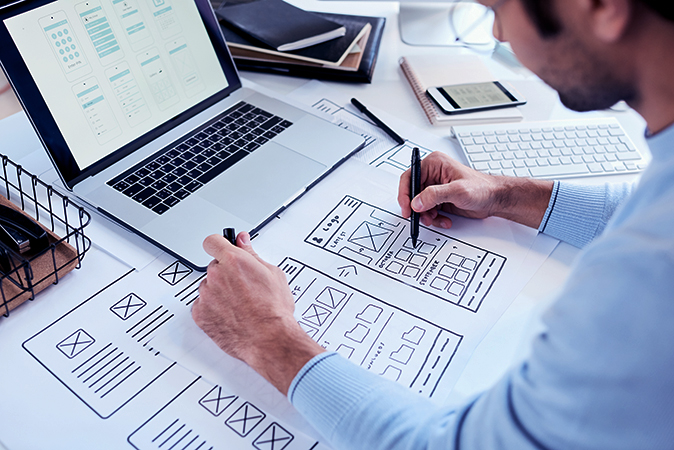 Close-up of busy designer sitting at table with laptop and drawing icons placement for ui design