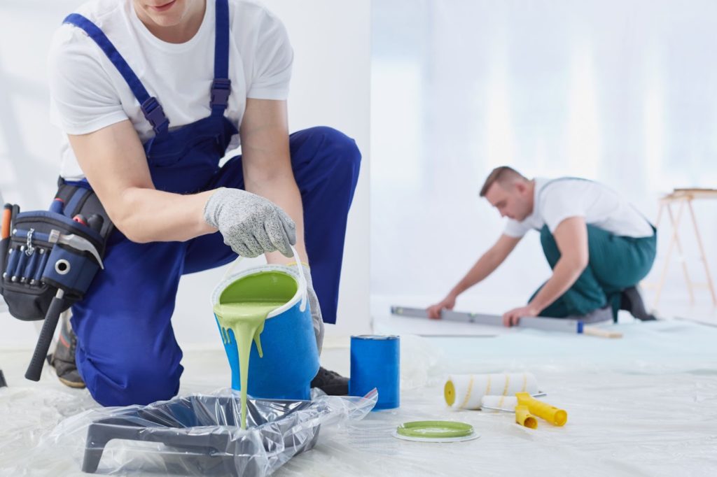 Two men painting on a white floor with paintbrushes and cans of paint nearby.