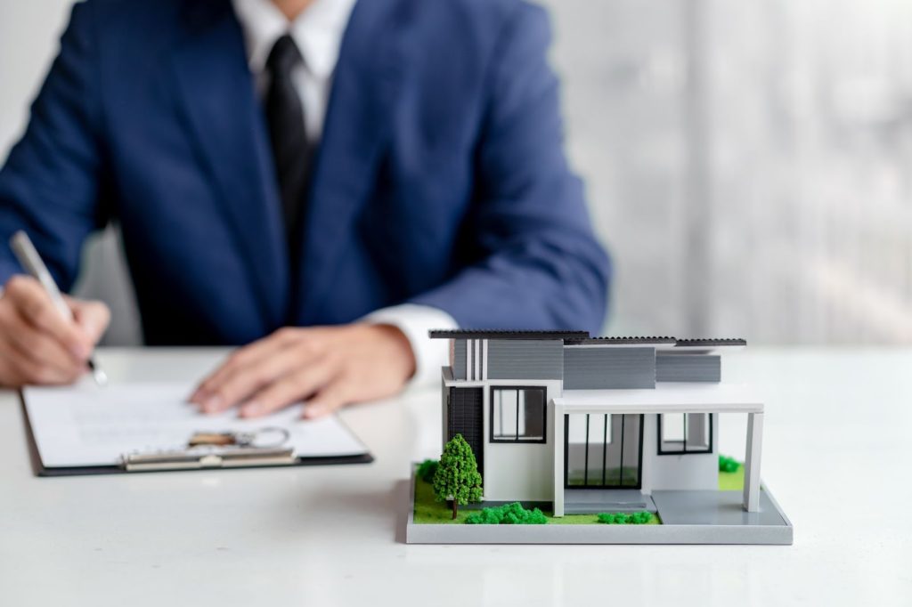 A businessman in a suit writes on paper with a model house, symbolizing real estate or construction plans.