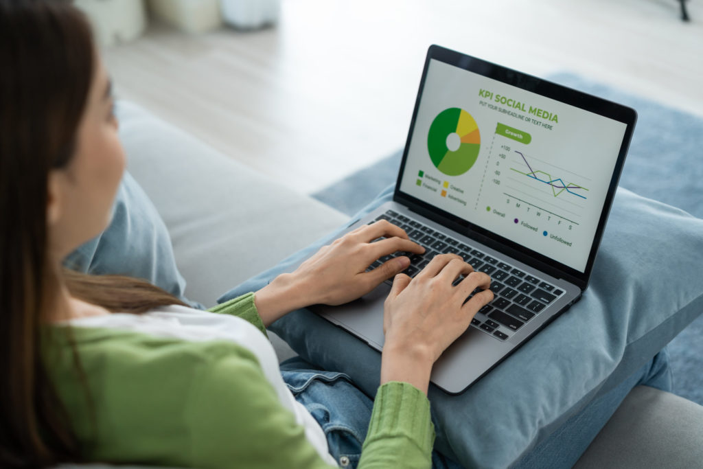 Rear view of Attractive beautiful business woman working from home. Employee office worker sitting on sofa in living room, using laptop computer typing on keyboard checking social media KPIs in house.