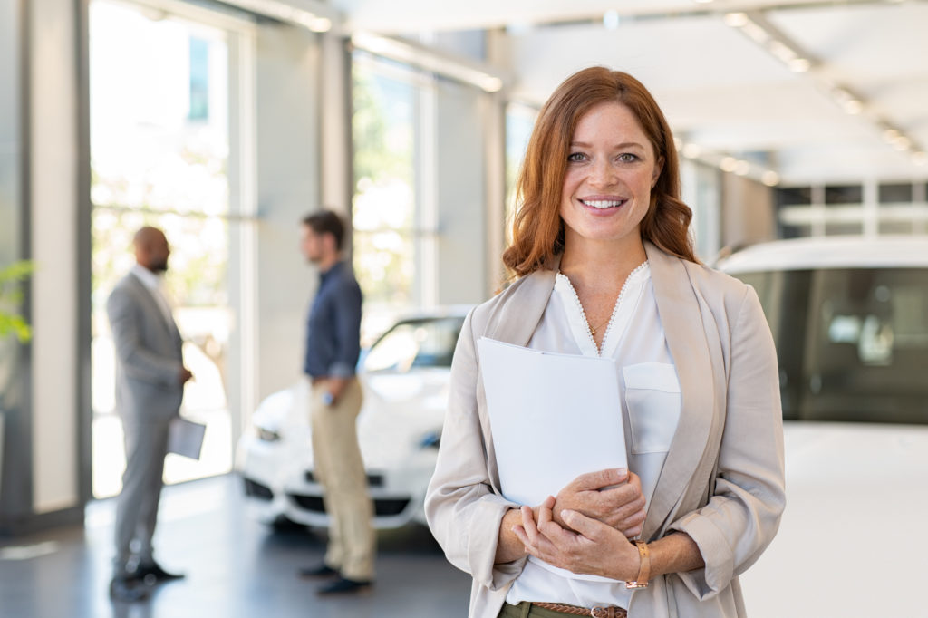 Smiling saleswoman holding document while looking at camera at car showroom. Young cardealer holding clipboard in automobile showroom. Professional confident sales person working in modern car dealership.