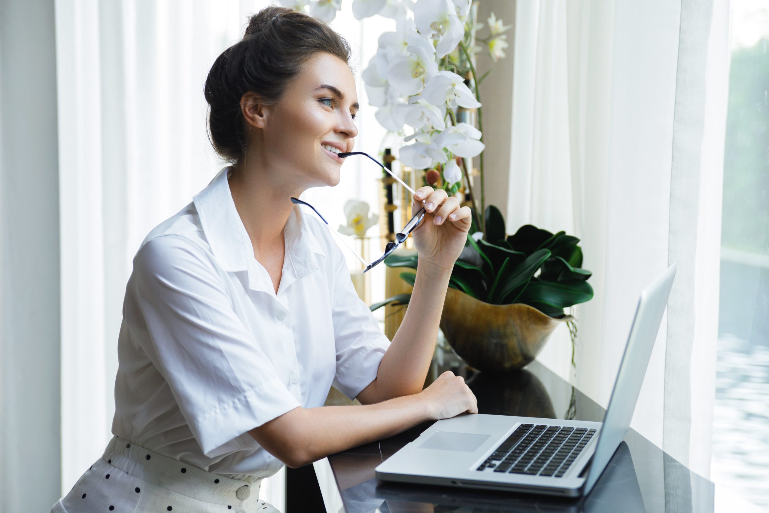 a woman sitting at a desk with a laptop and glasses in her mouth