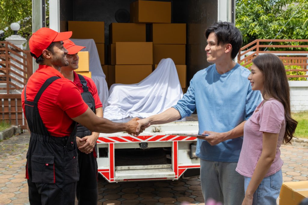 Two movers in red uniforms shaking hands with a young couple near an open moving truck loaded with furniture.