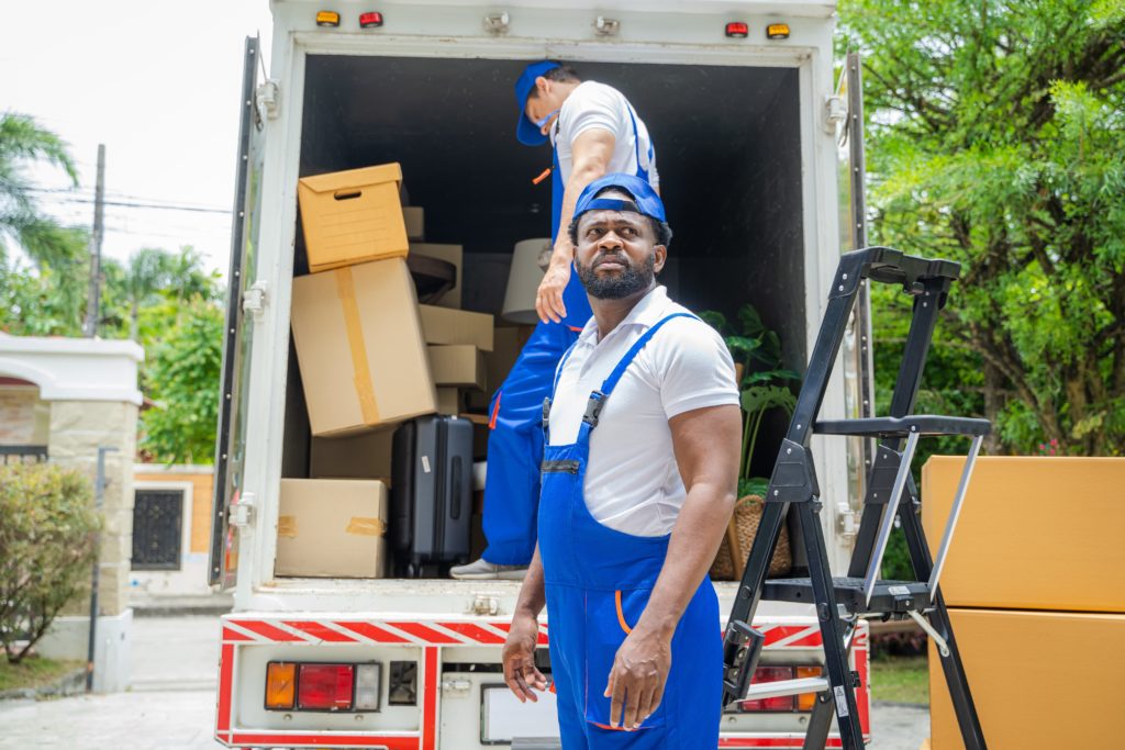 Two movers in blue uniforms unloading a truck filled with boxes and furniture in a residential driveway. .